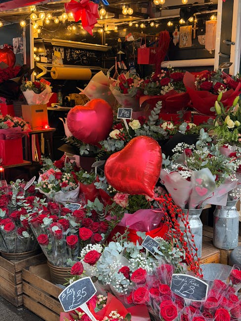 A vibrant display of floral arrangements featuring red roses, white flowers, and greenery, set among red heart-shaped helium balloons at a flower stall. The bouquet arrangements are wrapped in various
