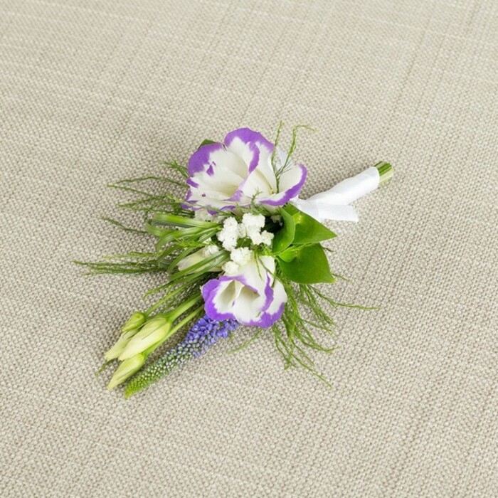 This image shows a thoughtfully arranged floral boutonniere placed on a softly textured, light beige fabric that resembles natural linen, its neutral warmth giving the flowers a calm, elegant stage. Dominating the composition are two open lisianthus blooms, their white petals unfolding in generous, ruffled layers, each edge kissed with an intense, velvety purple that creates a striking, bi-coloured ring around the centre. Between and around them sit clusters of very small, snow-white filler flowers-similar to baby's breath-adding a misty, romantic softness that lightens the overall look. Cutting through this gentle haze is a slim, upright spike of deep purple, bell-like florets that look like grape hyacinth, lending a distinctive texture and a more saturated note of colour, almost like a jewelled accent in the middle of the design. Two pale green buds rest nearby, still closed and smooth, suggesting blooms that will open later, echoing the hopeful feeling of a wedding morning somewhere in Sipson, perhaps just before heading to a local registry office or intimate venue. Surrounding everything is a mix of fine, feathery greenery and slightly broader leaves; the feathery sprigs, likely asparagus fern, contribute light, airy movement, while the deeper greens ground the boutonniere and frame the focal flowers. At the base, the exposed stems are neatly trimmed and bound together with a sleek white satin ribbon that encircles them tightly, finished in a small, tidy knot or bow, ready to sit securely on a suit lapel. The overall effect is one of classic, considered elegance with a whisper of countryside charm, the kind of handcrafted buttonhole a Sipson florist would prepare for a groom, best man, or prom guest wanting something refined yet gently expressive of the day's emotion.