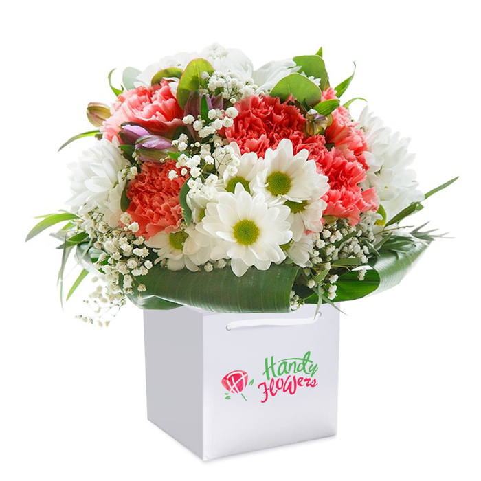Upright and indulgently full, this stunning bouquet captures the creative spirit of Sipson floristry, artfully displayed in a crisp, square white gift bag. The interplay of colour is striking: coral, peach, and red carnations-known for their ruffled, multi-layered petals-build a textured foundation teeming with warmth and energy. Bright white daisies, each with a vivid lime-green center, add lively contrast, their simplicity balancing the bouquet's richness. Sprigs of baby's breath weave gently between the larger blooms, bringing a gossamer lightness that suggests the serenity of the parks bordering Sipson. The arrangement is further deepened by a few closed, purple-tinged buds emerging discreetly on the left, introducing an element of anticipation. Glossy, deep green leaves wrap around the base, fanning out in bold, natural arcs that create a lush cradle for the blossoms-a testament to skilled local hands. The entire bouquet sits securely in a sleek gift bag equipped with smooth white ribbon handles for easy carrying, perfect for discreet delivery around Sipson, from Holloway Lane to the heart of the village. The image is shot against a flawless white background, highlighting the bouquet's vibrant hues and professional composition. Ideal for birthdays, anniversaries, or sending a heartfelt surprise, this arrangement epitomizes the freshness, elegance, and care that define flower delivery in Sipson.
