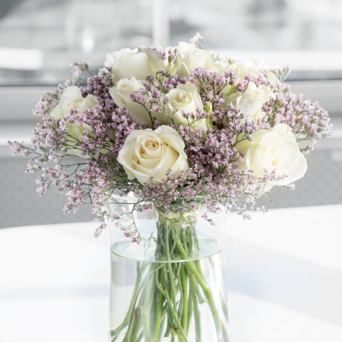A refined, contemplative bouquet fills the frame, made up of generous white roses and soft lavender filler flowers, arranged with a sympathetic touch in a clear glass vase. About a dozen roses are visible, each bloom open and full, their off-white petals shading gently into ivory at the centers. The petals curl and overlap in delicate spirals, creating a sense of depth and quiet luxury, while their smooth, almost satiny surfaces catch the light in a muted sheen. Interwoven among these central blooms are numerous sprigs of fine, branching statice, their tiny blossoms in pale lilac, lavender pink, and occasional soft mauve. These smaller flowers form a gentle, cloud-like surround, softening the structure of the bouquet and lending it an airy, almost meditative quality. The stems of both roses and filler are gathered cleanly into a tall cylindrical vase, the transparent glass revealing straight green stems descending into clear water that glints softly. The arrangement sits on a pristine white tabletop, which mirrors a subtle glow beneath the vase. Behind it, the background fades into a smooth, light blur, echoing the calm interiors of a quiet Sipson home or a small, sunlit room overlooking the local fields. The lighting is natural and diffused, as if filtered through a window on a mild morning, highlighting the bouquet's gentle contrasts without any harsh shadows. This image conveys the type of arrangement a Sipson florist might create for a funeral service, a condolence gift to a grieving family, or a simple vase to bring a touch of peace to a sideboard or bedside table, offering comfort through its understated beauty.
