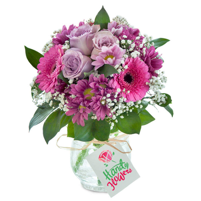 Taking inspiration from Sipson's friendly charm, this meticulously assembled bouquet radiates positivity and heartfelt sentiment from the first glance. At its core, pale lavender roses unfurl with a quiet grace, hinting at their soft, sweet perfume-reminiscent of early morning walks around Sipson Village Green. Large, striking Gerbera daisies in a brilliant fuchsia hue add a joyful focus, their cheerful faces and deep, expressive centres brightening the surrounding blooms. Threaded around them, magenta and plum chrysanthemums curl and cluster, their many-layered petals creating subtle shadows and texture throughout the design. Wisps of white baby's breath float between the more vivid flowers like tiny clouds, lifting and lightening the entire arrangement. Framing these vibrant blooms, lush deep green leaves peek through-a gentle nod to the hedgerows and gardens that are so much a part of Sipson's landscape. All these flowers rest securely in a rounded clear glass vase with a beautiful crackled finish, a unique touch that adds rustic character. Around the vase's neck is tied a strand of natural raffia, holding a plain white gift tag, ready for a handwritten message-a hallmark of local flower gifting in the Sipson area. This bouquet is an ideal choice for anyone wishing to deliver a sincere expression of joy, gratitude, or celebration, whether for birthdays, anniversaries, or those simple yet meaningful moments that brighten everyday life in Sipson and the surrounding neighbourhoods.