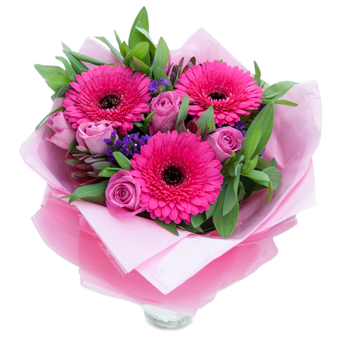 I see this arrangement as a graceful moment captured in pinks and purples: three bold, fuchsia Gerbera daisies command attention with their wide, layered petals and rich, dark centres, while a circle of lavender-tinged roses surrounds them, each rosebud held in a poised, almost secretive curl that promises unfolding softness. Little clusters of purple statice nestle into the gaps, their papery florets adding a subtle, rustic texture reminiscent of hedgerow blooms after a spring rain. The greens - broad, slightly glossy leaves resembling salal or eucalyptus - give the bouquet a structured, botanical backbone and a faintly aromatic, resinous note when handled. The flowers are wrapped for gifting in a soft, matte pale pink tissue that forms a gentle bed, topped by a translucent fuchsia-pink cellophane with a delicate crinkle that catches light and deepens the pink theme without overpowering the flowers themselves; the wrapping is carefully folded and finished, hinting at artisan hands. At the base, the rim of a clear glass vase is visible, showing cut stems and fresh water and underscoring the arrangement's immediacy and suitability for display. In the kind of soft morning or late-afternoon light you might find near Sipson Village or along the Bath Road, the colours glow warmly; the roses offer a faint perfume, the gerberas a clean, green nuance, and the overall effect is quietly celebratory - an elegant choice for a mother on Mother's Day, a close friend's birthday, or an anniversary where you want to convey both affection and thoughtful restraint.