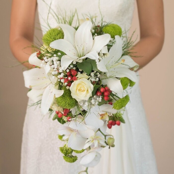 Against the gentle blur of a traditional ivory wedding gown, a joyful bride in Sipson tenderly holds a magnificent cascading bouquet-a testament to fine local floristry and inspired wedding artistry. Towering at the heart of the arrangement, several exquisite white oriental lilies feature broad, trumpet-like petals touched with subtle russet freckles, immediately drawing the viewer's eye. A central, tightly spiraled rose in soft cream adds a gentle, familiar romance. Alternating with the floral focal points, velvety lime-green mums pop with fresh vibrancy, their rounded forms lending texture and a modern accent reminiscent of local springtime walks along Sipson Lane. Interspersed throughout are clouds of dainty white baby's breath, their light, cloudlike blossoms infusing the arrangement with gentle movement and volume. Red hypericum berries, like tiny jewels, are peppered throughout, providing festive highlights that enliven the mostly white and green palette. The bouquet's signature feature-the graceful, cascading white Phalaenopsis orchids-tumble downward with sheer elegance, giving the ensemble its classic waterfall silhouette. Embellished with slender, undulating wisps of green and glossy foliage, the bouquet feels freshly gathered from the Sipson countryside. The bride's delicate lace sleeves and gentle grip on the bouquet echo the theme of romantic hope and anticipation, while expert composition ensures that each element is tastefully balanced. Perfect for spring and summer weddings in Sipson, this bouquet reflects both traditional bridal beauty and modern design sensibility, as cherished by clients of Flower Delivery Sipson.