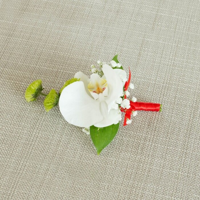 This image shows a meticulously crafted boutonniere arranged on a pale beige, linen-like fabric whose woven texture brings to mind natural, understated table linens in a Sipson country pub set for a wedding breakfast. Center stage is a flawless white Phalaenopsis orchid bloom, its generous petals unfolding with a soft, satin sheen. The heart of the orchid reveals a nuanced blend of buttery yellow and fine reddish-orange markings, drawing the eye inward and lending the piece artistic detail. Surrounding the orchid, delicate sprays of white baby's breath form a gentle halo, each tiny flower offering a whisper-light touch that softens the strong, sculptural presence of the central bloom. To the left, two rounded lime-green chrysanthemum or green trick-style pom-pom flowers introduce vibrant contrast, their tight, spiky texture creating a lively rhythm that feels contemporary yet playful, much like the energy of guests arriving for a ceremony in nearby Harmondsworth. Underneath, a broad, lush green leaf cradles the arrangement, its surface providing both structure and a rich, botanical backdrop that highlights the white petals. A smaller, darker green leaf peeks out near the stems, adding extra dimension. The stems are cleanly and securely wrapped in an intense crimson red satin ribbon, the fabric wound in precise, even coils and finished with a compact bow or knot that adds a polished, festive flourish. The neutral, slightly coarse fabric beneath the boutonniere ensures that each colour-the crisp white, vivid green, and striking red-stands out clearly. Overall, the accessory radiates elegant formality and quiet joy, perfectly suited to a groom, best man, or guest at a refined Sipson wedding or formal celebration nearby.