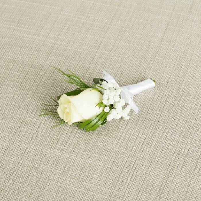 The image captures an elegant ivory rose boutonniere resting on a soft, neutral backdrop, as if waiting to be pinned just before guests arrive at a Sipson wedding venue. The background fabric is a light beige with a gentle diagonal weave, reminiscent of fine linen or natural cotton, its subtle texture lending a relaxed, organic feel. Placed diagonally across this canvas is a single rosebud in creamy white, its petals still held close yet clearly on the brink of opening, creating a sense of quiet anticipation. The rose's surface appears velvety and smooth, with a faint hint of green at the base where the sepals cradle the bloom, evidence of its freshness. Clustered to one side and tucked behind the rose are delicate sprays of white baby's breath, each tiny flower like a soft dot of foam, adding volume without overpowering the simplicity of the design. Surrounding both rose and gypsophila, slender dark green leaves and a few feathery, fern-like sprigs stretch out just enough to frame the central flower, giving structure and contrast while maintaining a clean, classic silhouette. The stems of the arrangement are neatly aligned and wrapped in a crisp white satin ribbon, which has been tied into a small, refined bow positioned just below the head of the boutonniere. The ribbon catches the light with a gentle sheen, hinting at the smoothness you would feel under your fingers. Soft, diffused lighting from above caresses the petals and foliage, revealing their form and subtle shadows without harshness. Created with the care of a local florist familiar with ceremonies across Sipson and nearby Harmondsworth, this boutonniere conveys timeless elegance, ideal for grooms, ushers, or fathers of the couple seeking a traditional, understated floral accent.