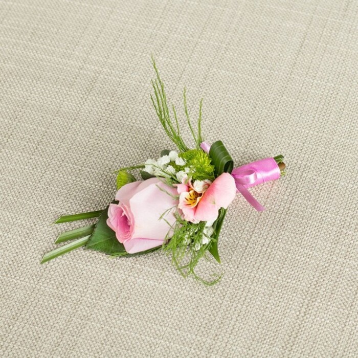Captured on a neutral, subtly textured linen-something reminiscent of Sipson's traditional market stalls-a refined floral boutonniere sits poised as if waiting for its moment to shine at a special village event. The focal point is a tightly furled, blush pink rosebud, its petals hinting at the promise of unfurling beauty. Flanking the rose, a softly ruffled Alstroemeria in tones of peach and pale pink introduces swirling patterns of subtle yellow with darker markings near the core, while fine wisps of baby's breath float between the larger blossoms, adding a breathy lightness to the arrangement. Petite green cushion mums lend a burst of fresh texture, balanced by feathery sprigs of green reminiscent of the foliage bordering Sipson Way. The boutonniere is framed with a rich variety of greenery: pointed grass blades fan upward, and broader, darker leaves create a natural cradle, their glossy surfaces occasionally folded to echo the careful work of a practiced hand. Encasing the stem binds, a wide dark green leaf is neatly wrapped and secured with a chic fuchsia pink satin ribbon, its bow gleaming amid the more matte textures, infusing the accessory with celebratory flair. The scene is illuminated by soft, even lighting that enhances every petal, leaf, and fiber of the linen background, ensuring each detail stands out vividly. Designed with both tradition and modern taste in mind, this enchanting piece is suitable for weddings at local venues or school proms held in the heart of Sipson, exuding sophistication and heartfelt care in every element.