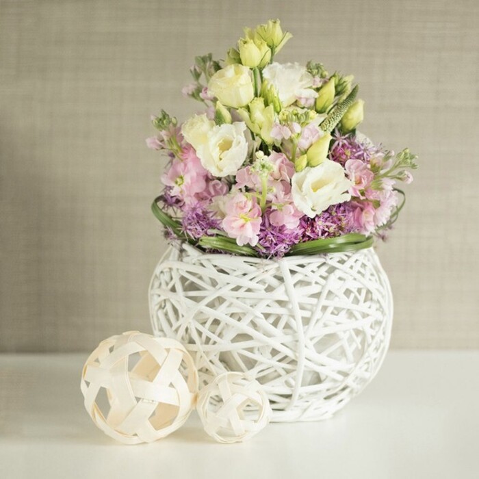 Amidst the quiet elegance of a softly lit Sipson interior, a beautifully lush floral ensemble unfolds atop a gleaming white surface. The bouquet radiates with creamy white and pale yellow lisianthus, each bloom displaying gently gathered, multi-layered petals that suggest the first signs of spring blossoming across local gardens. Vertical accents of pastel pink matthiola infuse height and gentle movement, while a cluster of vivid purple filler flowers forms a rich, tactile bed under the main blossoms, highlighted by a border of deep, fresh green leaves. The arrangement is expertly placed in a spherical white vase-its open, airy construction is reminiscent of finely woven painted rattan or delicate branches, combining a modern sensibility with the timeless beauty familiar to Sipson's florists. Flanking the display, two round decorative orbs, gently woven from natural rattan, echo both the vessel's form and the area's handcrafted aesthetics, often seen in local artisan markets. The soft, textured greige backdrop calls to mind the elegance of understated home décor in Sipson village, letting the florals' pastel colours and lavish shapes stand apart. Whether for a wedding breakfast in a Sipson venue or as inspiration for natural home styling, this arrangement embodies freshness, serenity, and a touch of artisanal charm, inviting peaceful reflection and understated celebration.