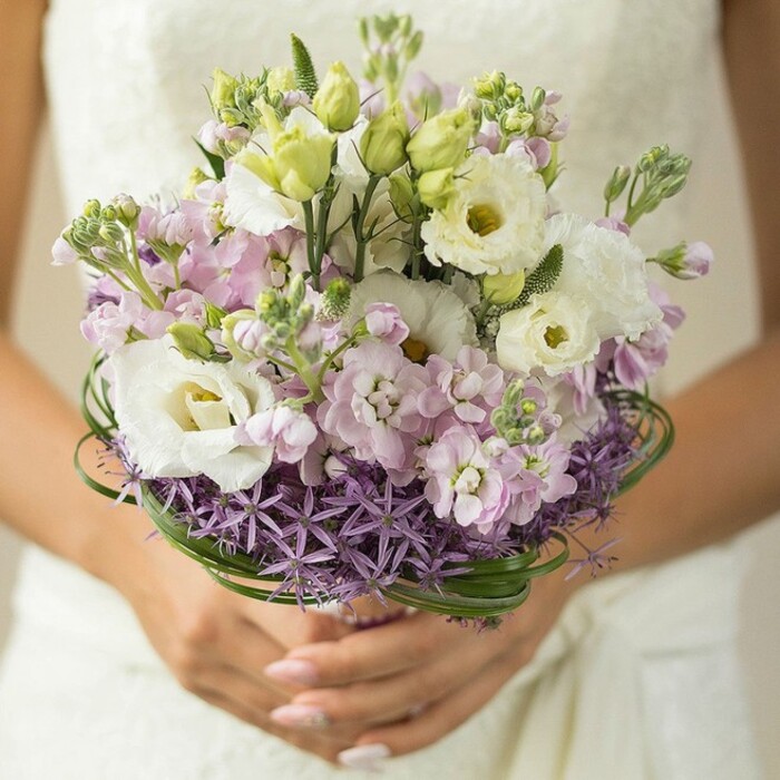 A bride in Sipson holds an exquisitely balanced bouquet close to her, the scene illuminated by natural light streaming through a softly blurred background, revealing a glimpse of her ivory lace wedding gown. The bouquet takes center stage-a lush, rounded composition anchored by elegant white Lisianthus flowers, their gently ruffled petals catching the light and drawing the eye inward to subtle yellow-green centers. Nestled amidst these are clusters of pastel pink and lilac stock flowers, rich in texture and warmth, their multi-petaled forms lending a romantic, dreamlike quality. Slender, fresh Lisianthus buds-pale green and full of anticipation-peek between the blossoms, hinting at new beginnings. Encircling the lower tier of the bouquet, a collar of starburst purple Allium creates a unique flare; the spiky blooms impart an architectural strength that contrasts beautifully with the arrangement's otherwise soft palette. Artful rings of glossy, deep green bear grass wind gracefully just above this base, adding a tailored finish and highlighting the florist's expertise. The bouquet's wrapped stem is cupped by lightly manicured hands-nails painted in a gentle nude-that support the flowers with tender care. This floral design, ideal for a spring or summer event in Sipson, reflects both contemporary styling and timeless romanticism, inviting thoughts of ceremonies in peaceful local gardens or along shaded village roads. Every detail celebrates the careful touch and artistry found within Sipson's vibrant floral community, making this bouquet a true tribute to love and the beauty of the season.