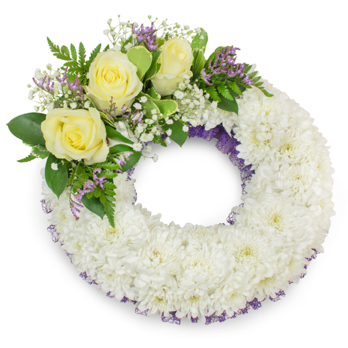 Presented against a pristine white background, this circular sympathy wreath is built from hundreds of small white chrysanthemums tightly arranged to create a smooth, plush ring that reads with comforting solidity. The chrysanthemum heads, each with layers of delicate petals, are placed to form an almost tapestry-like surface; the white field is interrupted only by a fine lining of deep purple mesh or ribbon glimpsed along the inner and outer edges, which gives the wreath a refined contour and a hint of sombre colour. In the upper-left quadrant a carefully staged spray breaks the symmetry: three soft yellow roses act as the visual and emotive anchor, their multi-layered petals offering a velvet softness against the crisp chrysanthemum ground. Clusters of tiny white gypsophila float around the roses like gentle halos, while sprays of lavender-toned statice inject subtle contrast and a slightly papery texture. A selection of lush green foliage, mixing broad leaves and delicate fern fronds, frames the bouquet cluster and provides depth and movement to the design. The open centre remains clear, symbolically suggesting continuity and remembrance, and the even, soft lighting highlights petal edges and the natural sheen of the leaves without harsh shadows. As a thoughtfully composed memorial piece suited to services in and around Sipson - from nearby Bath Road chapels to community gatherings at Sipson Green - it reads as a respectful, classic tribute crafted with technical skill and heartfelt intention.