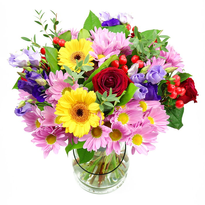 In this photograph, a lush and carefully curated bouquet sits in a clear glass vase, illuminated against an uncluttered white background that makes every detail stand out. The flowers look freshly arranged, as though they have just been prepared by a local Sipson florist for same-day delivery. At the core, deep red roses open in full, velvety blooms, their rich, wine-toned petals curling outward in elegant spirals that draw the eye inward. Flanking them are two bright yellow gerbera daisies with dark brown-orange centres, their bold, daisy-like faces adding a sense of joy and openness to the composition. Nestled around and between these star blooms are generous clusters of soft pink and light lavender daisies or chrysanthemums, each with fine, radiating petals and small golden centres that give the arrangement a feathery, cushiony texture. Purple and lilac lisianthus blossoms emerge gracefully on slender stems, their slightly ruffled, bell-shaped petals adding cool notes that temper the warmer reds and yellows. Scattered throughout, glossy red hypericum berries stand proudly on thin stalks, providing small, rounded accents that give the eye something surprising to discover. The bouquet is richly framed with greenery-broad, dark leaves give weight at the base, while airy, pale green sprigs reach outward, softening the edges and suggesting natural growth. Through the transparency of the vase, the neatly aligned green stems and clear water are visible, reinforcing a sense of freshness and care. The lighting appears even and natural, with gentle highlights on the petals and leaves, making the colours vibrant but true. The overall impression is of a celebratory, uplifting arrangement suited for birthdays, thank-you gestures, or to bring a touch of colour into a home near Sipson village, evoking both tenderness and cheerful good wishes.