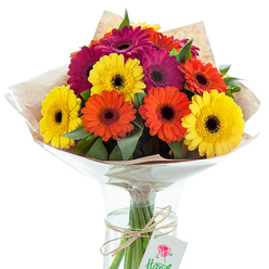 A dazzling hand-tied bouquet of Gerbera daisies is arranged with expert care in a clear glass vase, its vibrant display reminiscent of the cheerful blossoms seen at family gatherings along Sipson Lane. The composition brings together radiant yellow, fiery orange, and sumptuous deep magenta Gerberas, each marked by their signature dark, velvety centers, contrasting crisply with the brightness of their petals. The flowers sit nested among sprigs of supple green foliage, lending a layered texture and vibrant energy that mirrors the lush landscapes surrounding Sipson Recreation Ground. The bouquet is artfully wrapped: an inner sheath of lightly crinkled, translucent cellophane imparts a subtle sheen and preserves freshness, while an outer layer of earthy-hued brown kraft paper offers a rustic, tactile finish familiar to the bouquets crafted for local celebrations. Visible through the clear vase, each strong green stem is supported by fresh, cool water, helping the flowers keep their just-picked vibrance for delivery across Sipson and neighbourhoods like Heathrow Village. A length of natural twine fastens the stems with a decorative knot, suspending a minimal white tag with a delicate rose design-suggesting the bouquet is perfect for personal gifting. Soft natural light accentuates the abundance of colour, making this an uplifting and exuberant floral choice, whether to celebrate a milestone or simply to bring brightness into a home or workspace within the Sipson community.